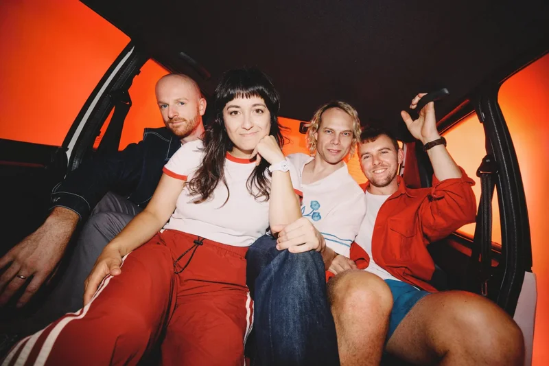 Picture of The Beths band members sitting in car interior against orange backdrop.