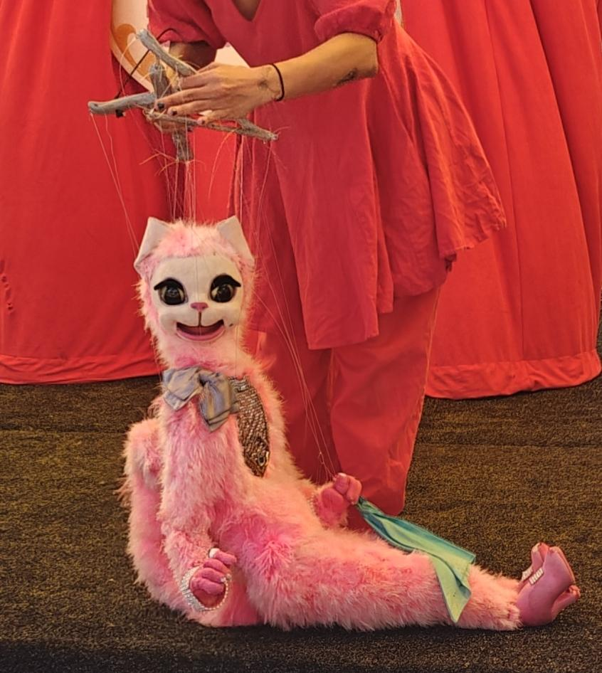 Pink cat marionette sitting on stage. Red background. Puppetteer also wearing red.