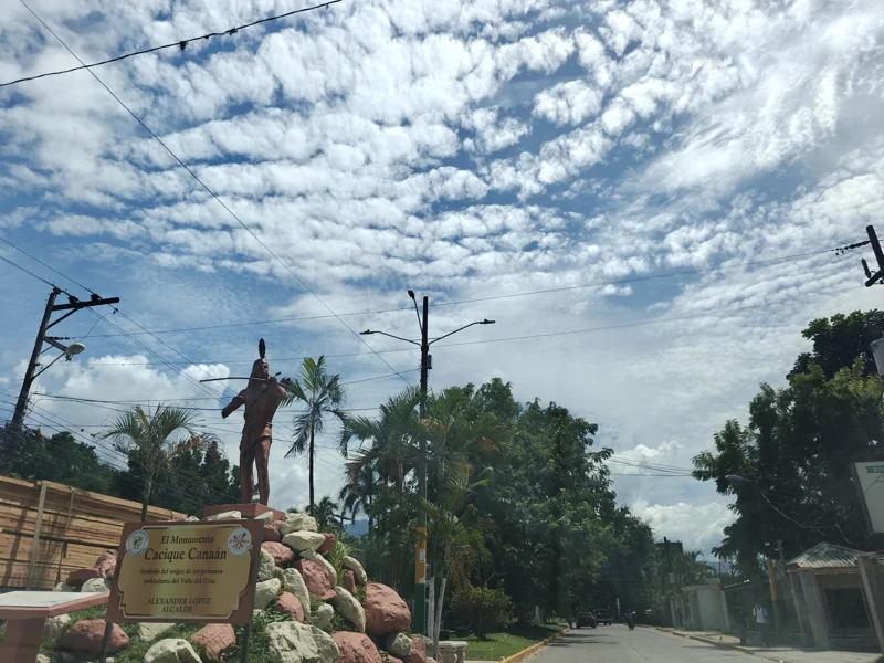Statue of native standing on rocks. Trees and powerlines. Vast clouds in the sky.