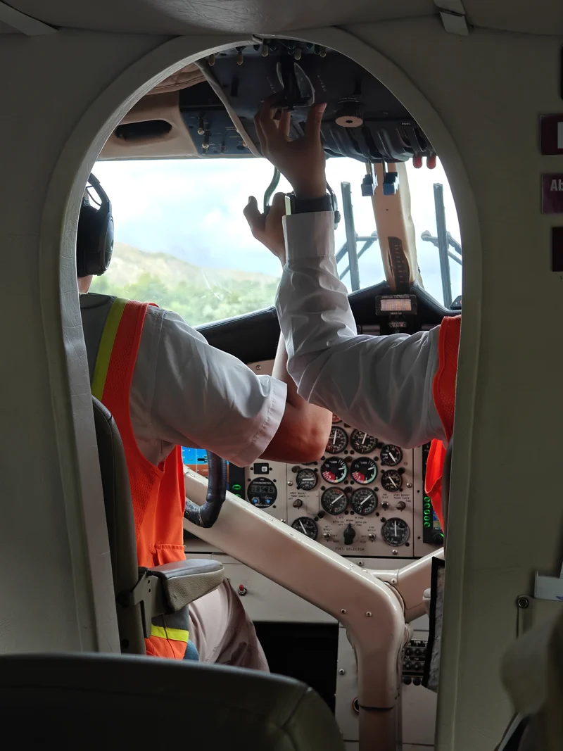 Open arched doorway to cockpit of small plane. Pilots are reaching upwards, and control panel has many dials.