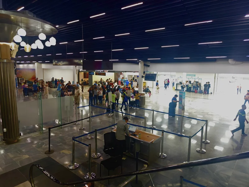 View from top of escalator toward groups of people at an airport. Three familiar faces are fading away.