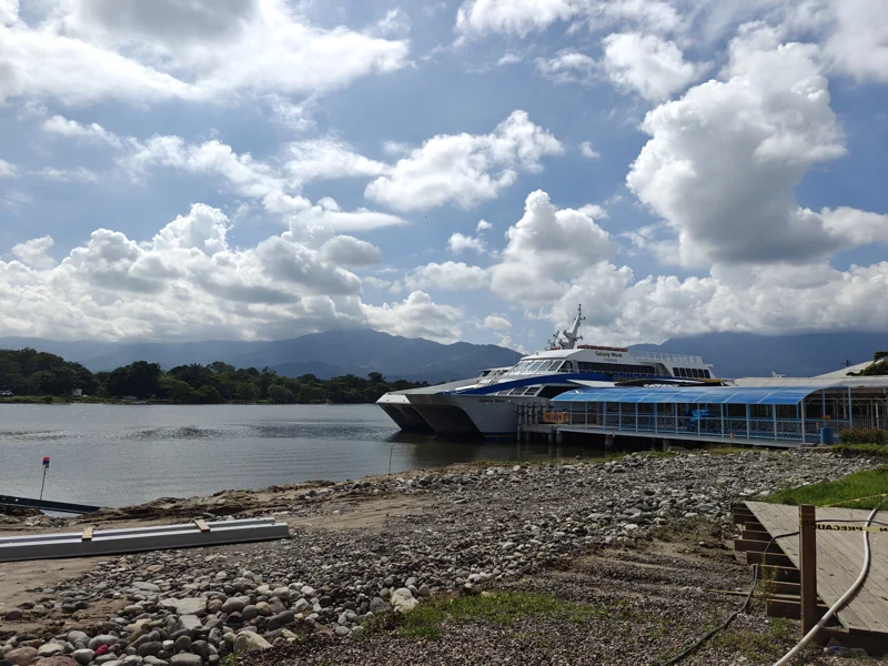 Rocky shoreline with dock leading out to a ferry boat on the water. Large puffy white clouds in the sky.