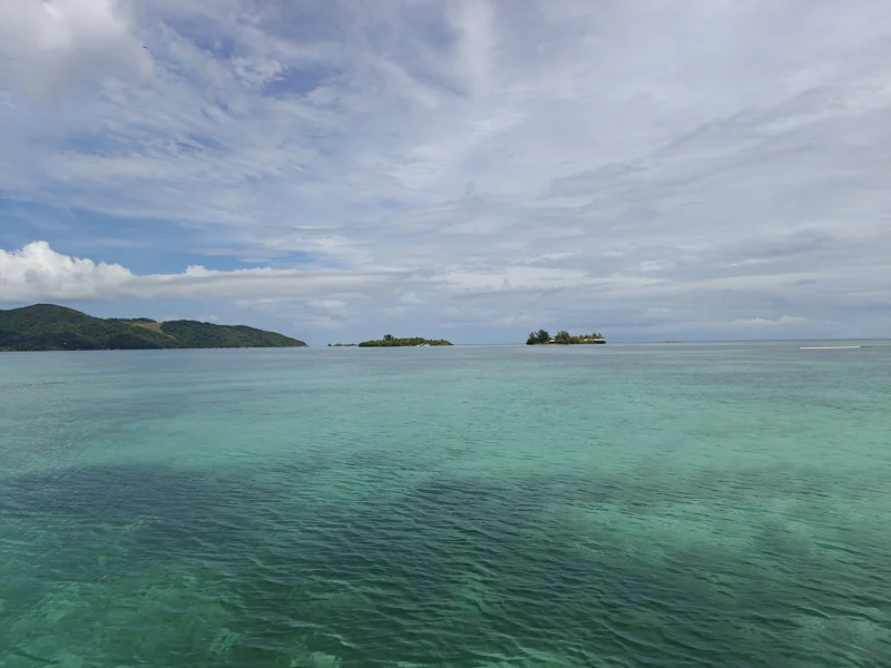 Extremely clear blue/green water and expansive sky. Island greenery in the distance.