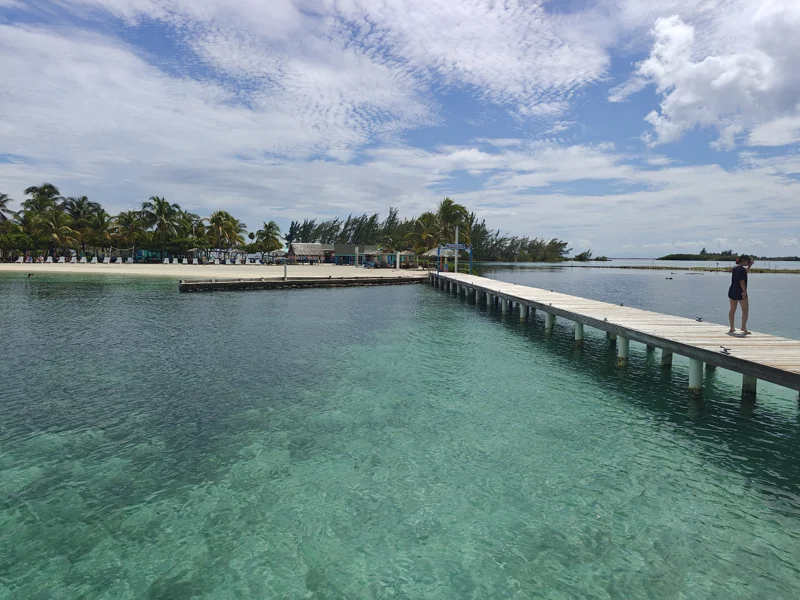 View from dock back towards beach. Water is crystal clear. Sky is expansive with white clouds.