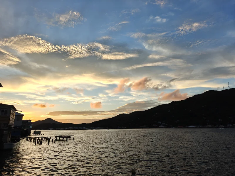 Shimmering water at dusk with watercolor clouds in the sky. Shadowy island beyond and houses to the left.