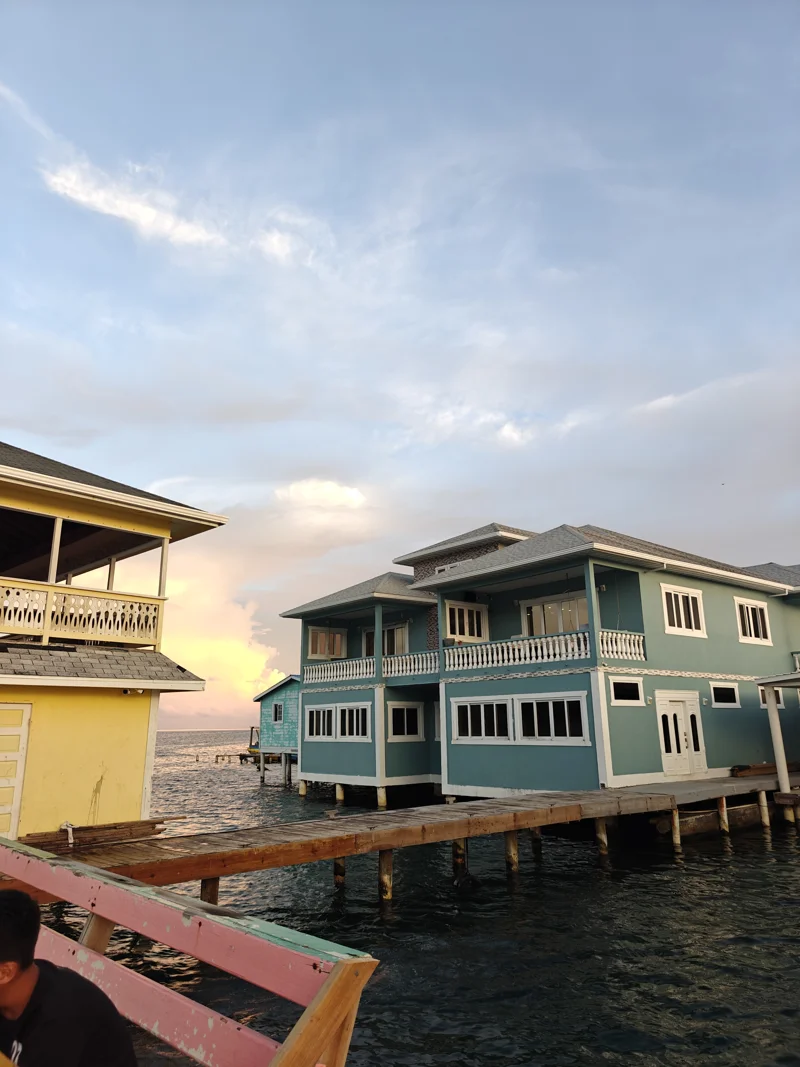 A green house built on the water next to a dock, leading to another yellow structure.