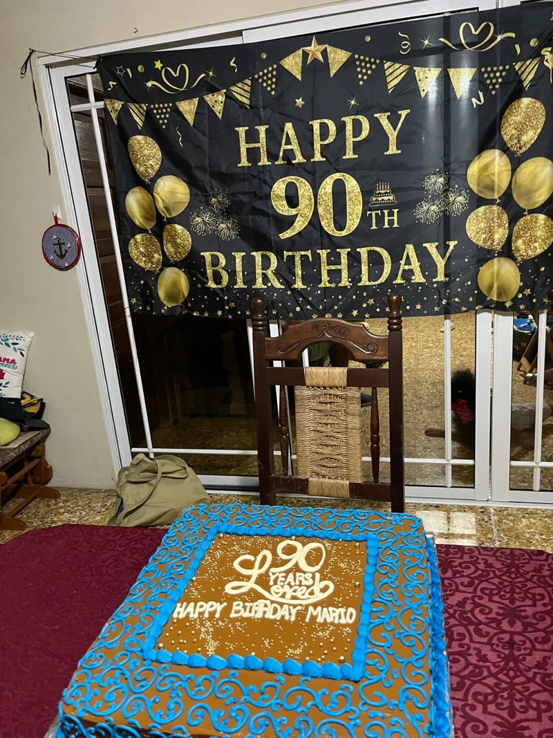 Big banner that says Happy 90th Birthday in gold lettering. An empty chair in front of a large cake with blue frosting. It says '90 Years of Love. Happy Birthday Mario'