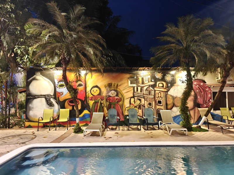 Pool with a mural beyond it painted in bright paint. Two palm trees next to several poolside chairs.