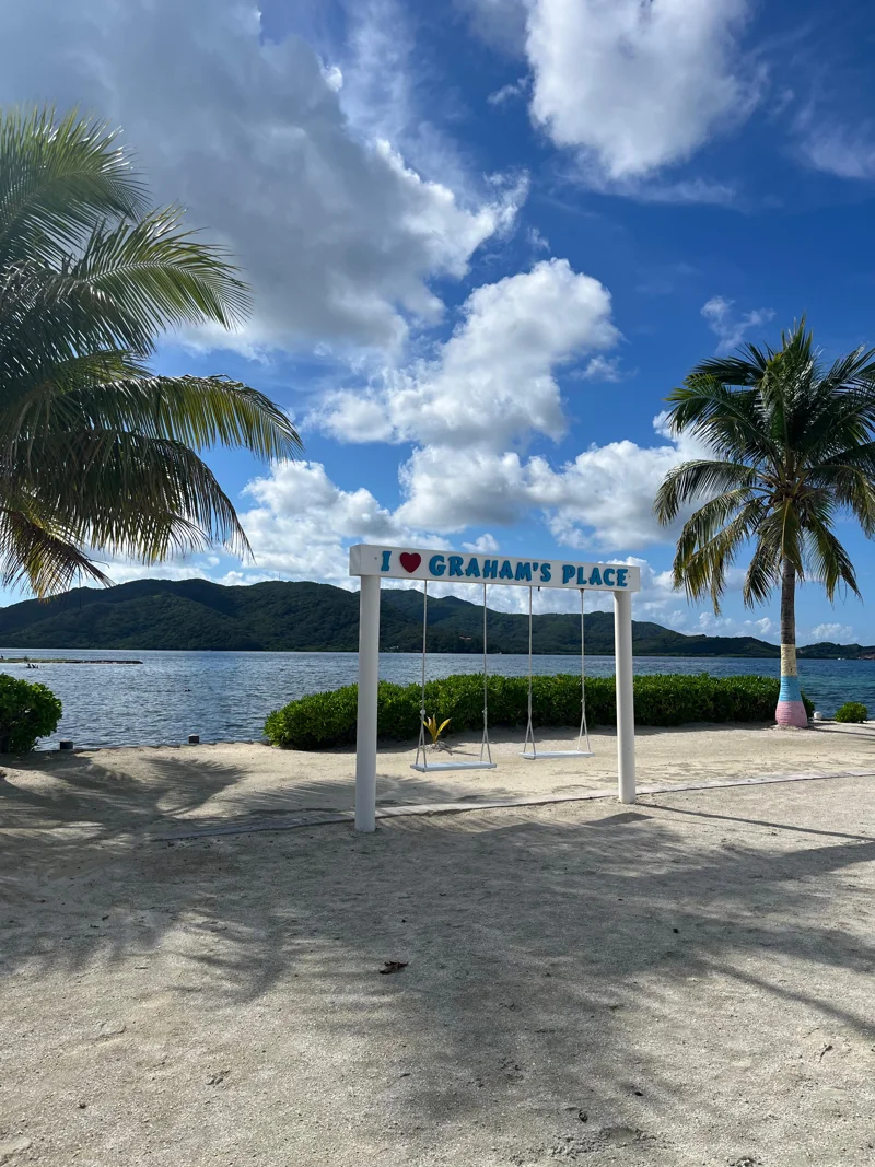 Sandy beach, with two swings hanging under wooden support structure that says I love Graham's Place. Palm trees, water, clouds.