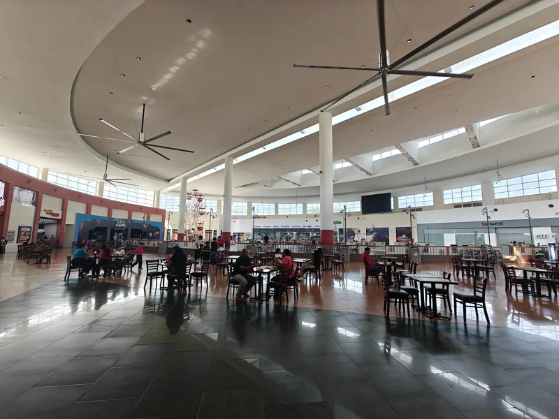 Wide open area with tables and seats. Some people eating. Large fans above.