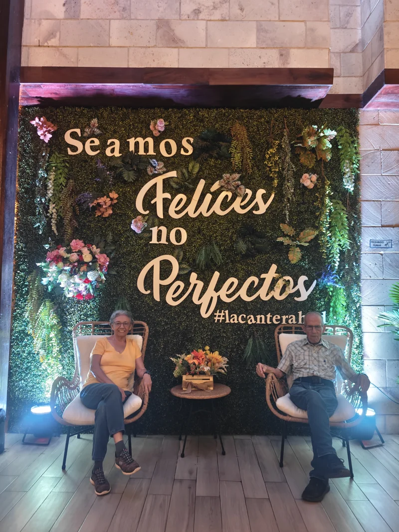 A wise couple sits in front of large signage, itself surrounded by greenery. The sign reads 'Seamos Felices no Perfectos'.