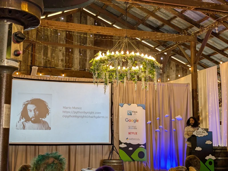 Stage inside a barn. On the screen there is a pictured of bearded man with disheveled hair and identifier that says 'Mario Munoz'. I'm standing on the stage on the right.