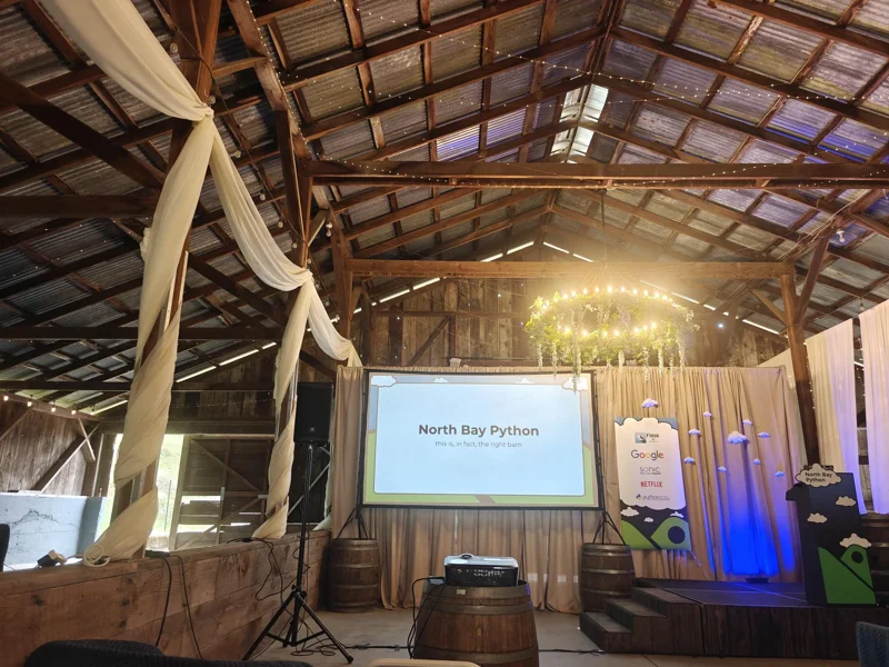 Stage inside a barn with wooden rafters. There is a screen that reads 'North Bay Python' and a small stage and lectern to the right.