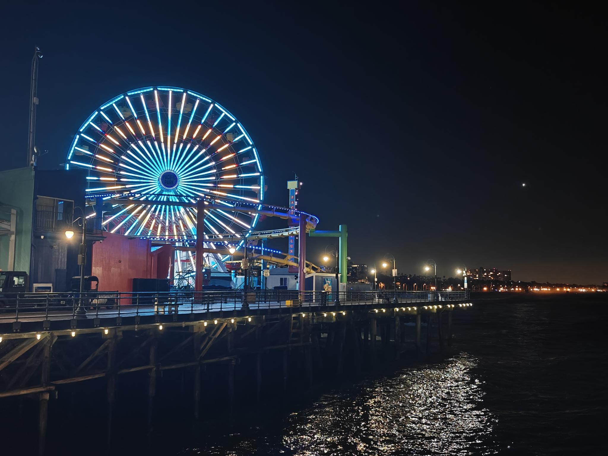 Santa monica pier at night. The ferris wheel is lit up with blue and yellow colors, making the shape of a heart. The lights reflect from the dark water below.