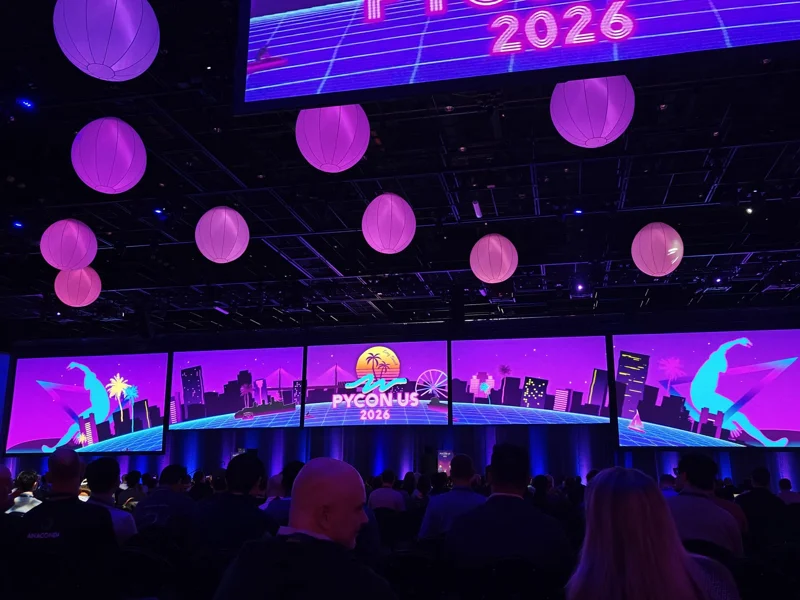 Dark conference room with screens across back wall with PyCon US 2026 logo and neon purple and pink theme. Pink orbs hang from the ceiling.