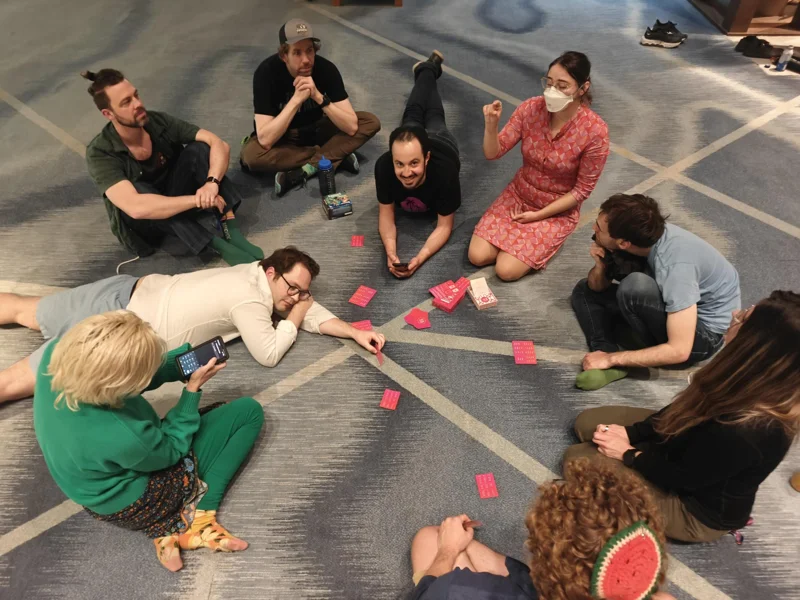 Shot from above. Nine people sit or lay on the floor with cards strewn in front of them.