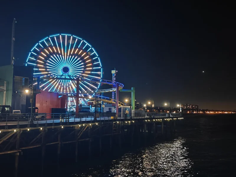 Ferris wheel on pier lit up with blue and yellow lights, making a heart. Below is dark water, reflecting light from the lamps on the pier.