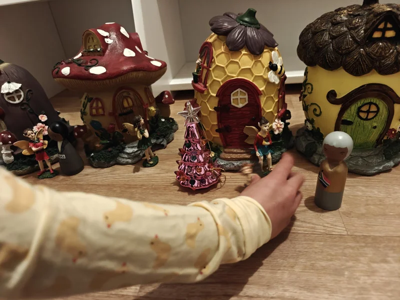 Hand reaching down holding ceramic fairy, next to four fairy houses of different colors. One resembles an acorn, one a honeycomb, and one a mushroom. Also, a wooden figure of Nelson Mandela in front of the acorn house.