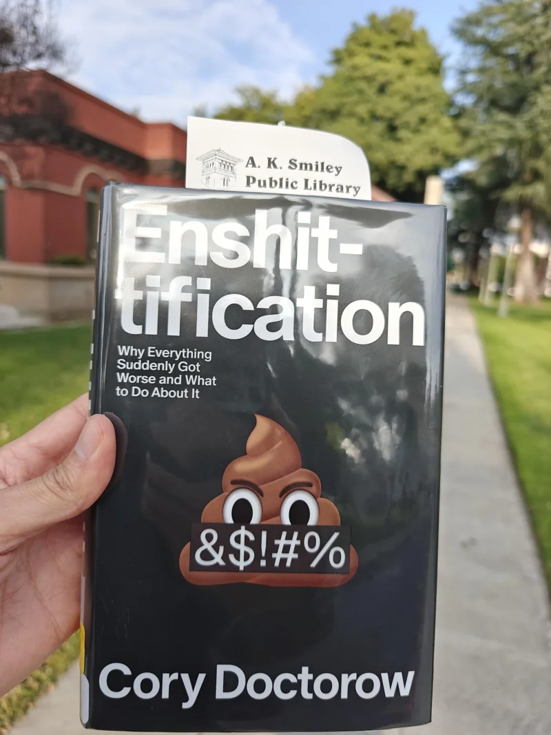 Cover of Enshittification by Cory Doctorow. Held up by a hand, with a library receipt sticking out. A blurred library and trees in the background.