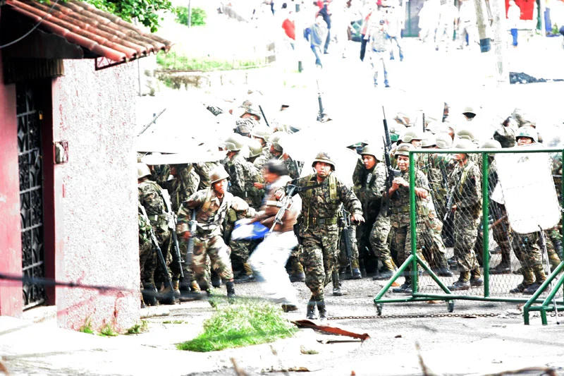 Camouflaged soldiers storming past a green fence toward a structure with clay tiled roof.