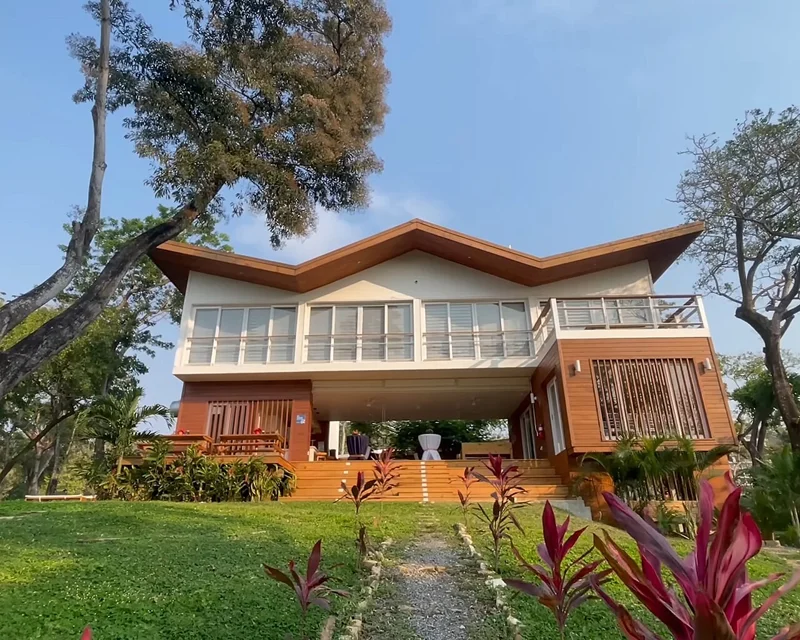 Green lawn with tropical red, leafy plants. Path leading up to wooden structure with stairs. The middle is hollow, with chairs and other furniture there. The second floor is white and has many windows on its face. The roof is zig zag. The sky is blue and trees surround the structure.
