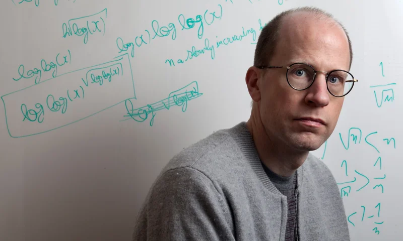 Man wearing glasses staring at camera. Behind him is a dry-erase board with equations written in green marker.