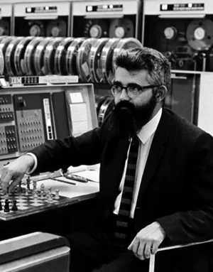 Black and white image of man with dark beard and glasses. Wearing suit with hand on a checkerboard with old computing equipment behind him.