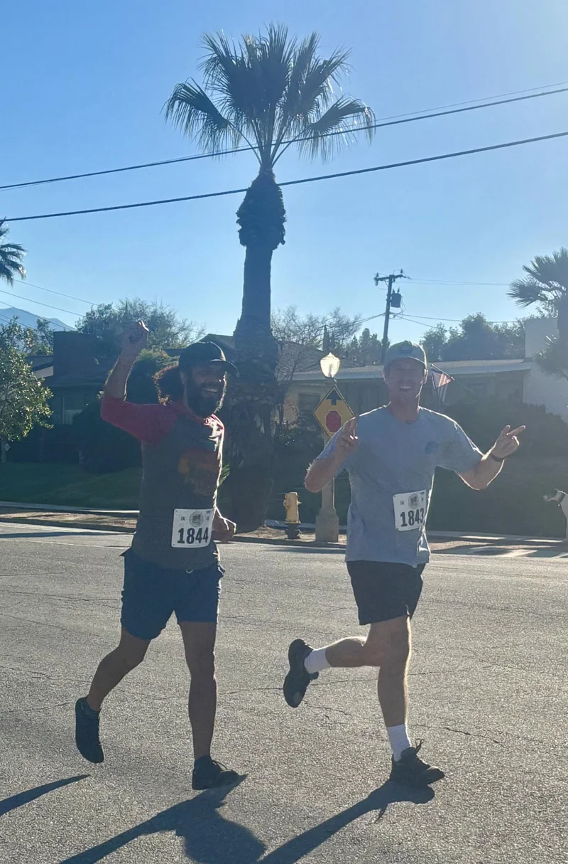 Two men running on a street with palm tree behind them. One is pumping a fist, the other is holding up peace signs with both hands.