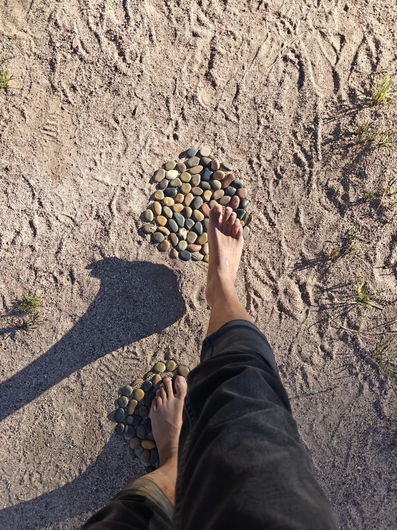 Legs and barefoot feet walking over small stones laid out in a circle atop of sand.